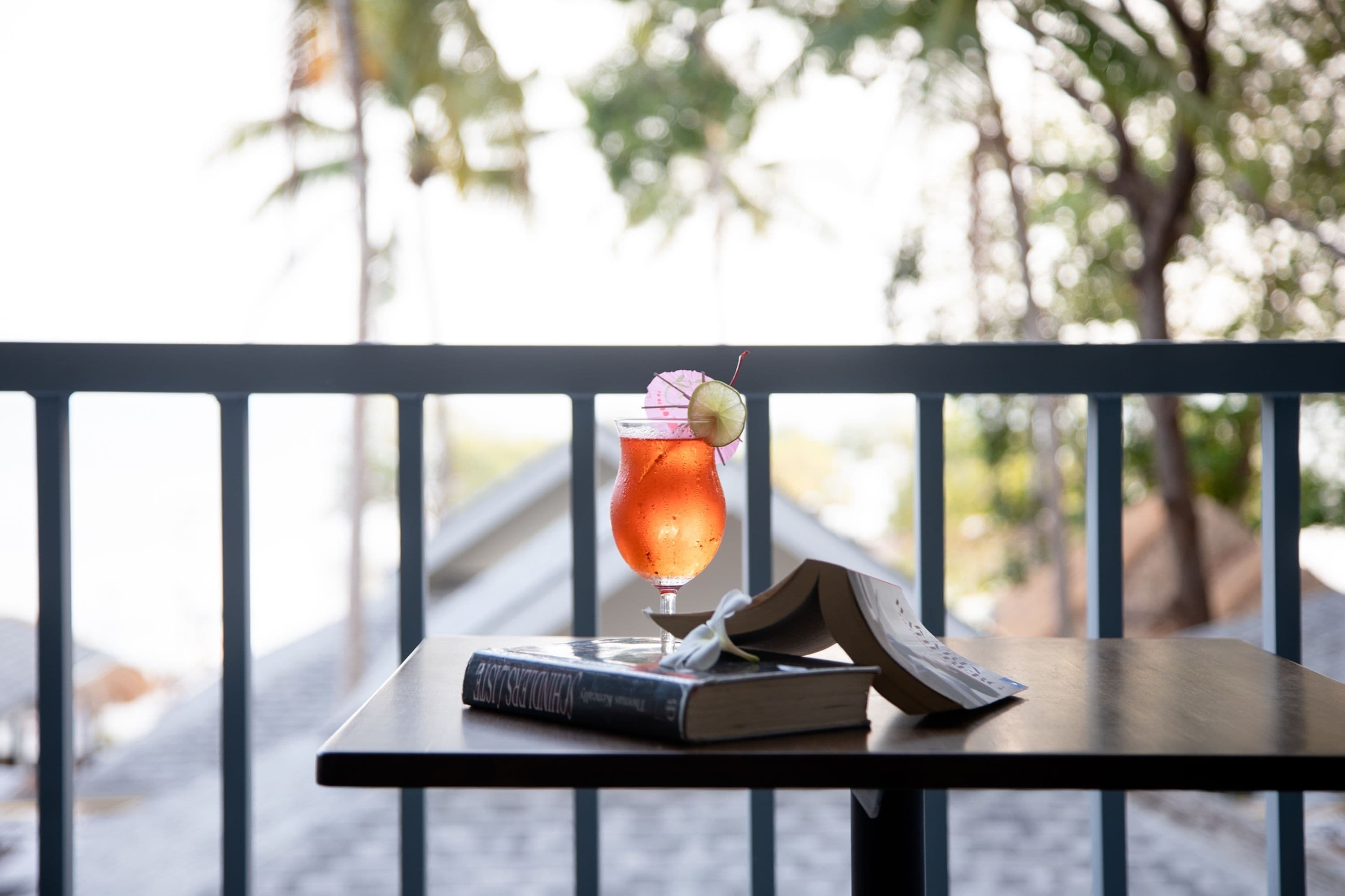 A table with a drink and a book, set against a railing with a blurred outdoor background.