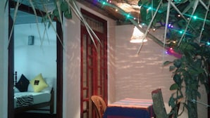 Ein Zimmer mit einem Bett, einem Tisch und einem Stuhl. Es gibt ein Fenster mit rotem Rahmen und eine Tür. Das Zimmer hat ein Strohdach und einige hängende Pflanzen.