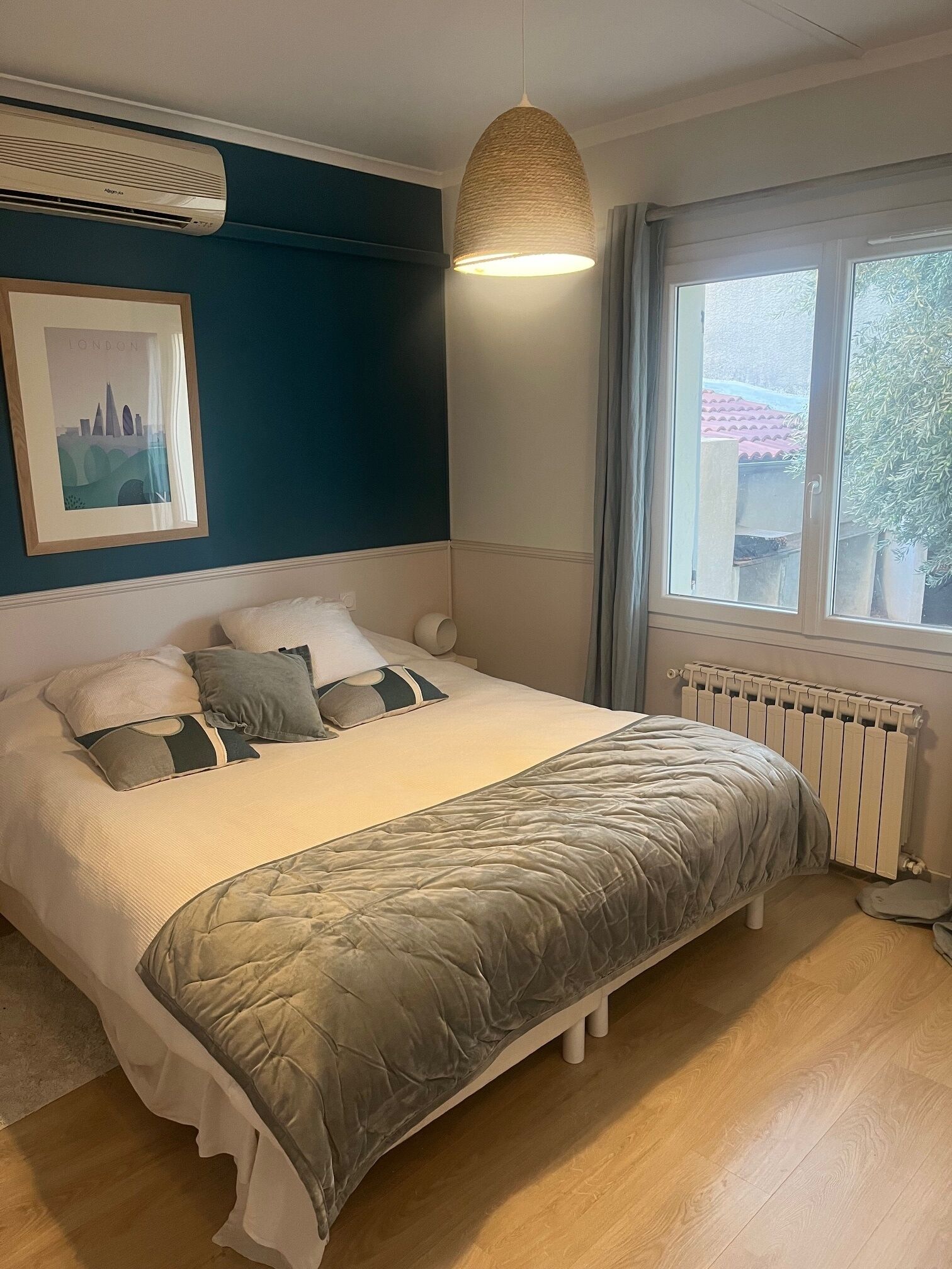 A bedroom with a bed, a hanging light fixture, a radiator, and a window with a view of the outside.