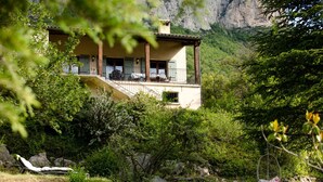 Um edifício de dois andares com varanda, cercado por vegetação exuberante e montanhas ao fundo.