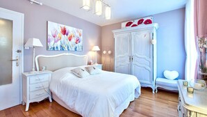 Um quarto com uma cama branca, uma mesa de cabeceira com abajur, um guarda-roupa e um quadro na parede.