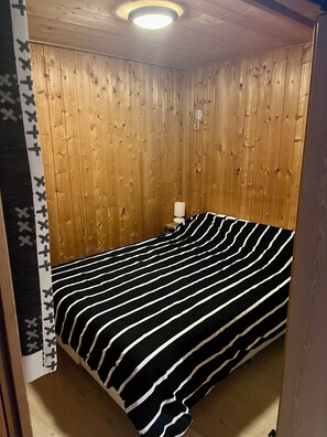 Ett litet rum med en enkel säng, väggar beklädda med träpaneler, ett svart- och vitrandigt sängöverkast och ett nattduksbord med en lampa.