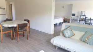 A spacious living area with a round dining table and chairs, a sofa with decorative pillows, a bed with a white duvet, and a small desk with a lamp. - 50m² studio with garden near Toulouse (Baziège)