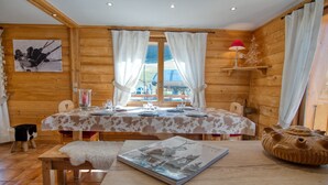 Interior de una cabaña de madera con una mesa de comedor preparada para cuatro personas, un cuadro en la pared y una ventana con cortinas.