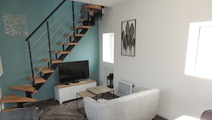 Ein modernes Wohnzimmer mit Fischgrätparkett, einem grauen Sofa, einem Flachbildfernseher auf einer Holzkonsole, einer Treppe mit schwarzen Geländern und einem Wandbild.
