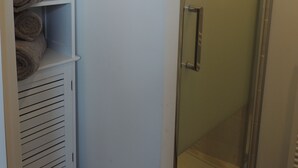 Eine Duschkabine mit Glas, eine weiße Tür, ein weißes Ablagefach mit Handtüchern und eine weiße Wand.