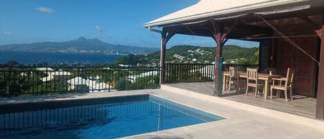 Ein Pool mit Blick auf das Meer und die Berge, eine überdachte Terrasse mit Stühlen und ein Gebäude mit weißem Dach.