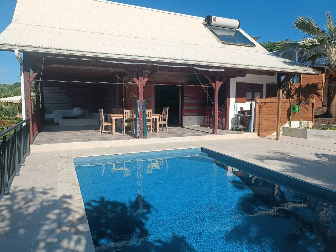 Une piscine avec un patio couvert, des meubles en bois et un palmier.