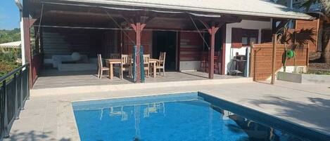 A swimming pool with a covered patio area, wooden furniture, and a palm tree.