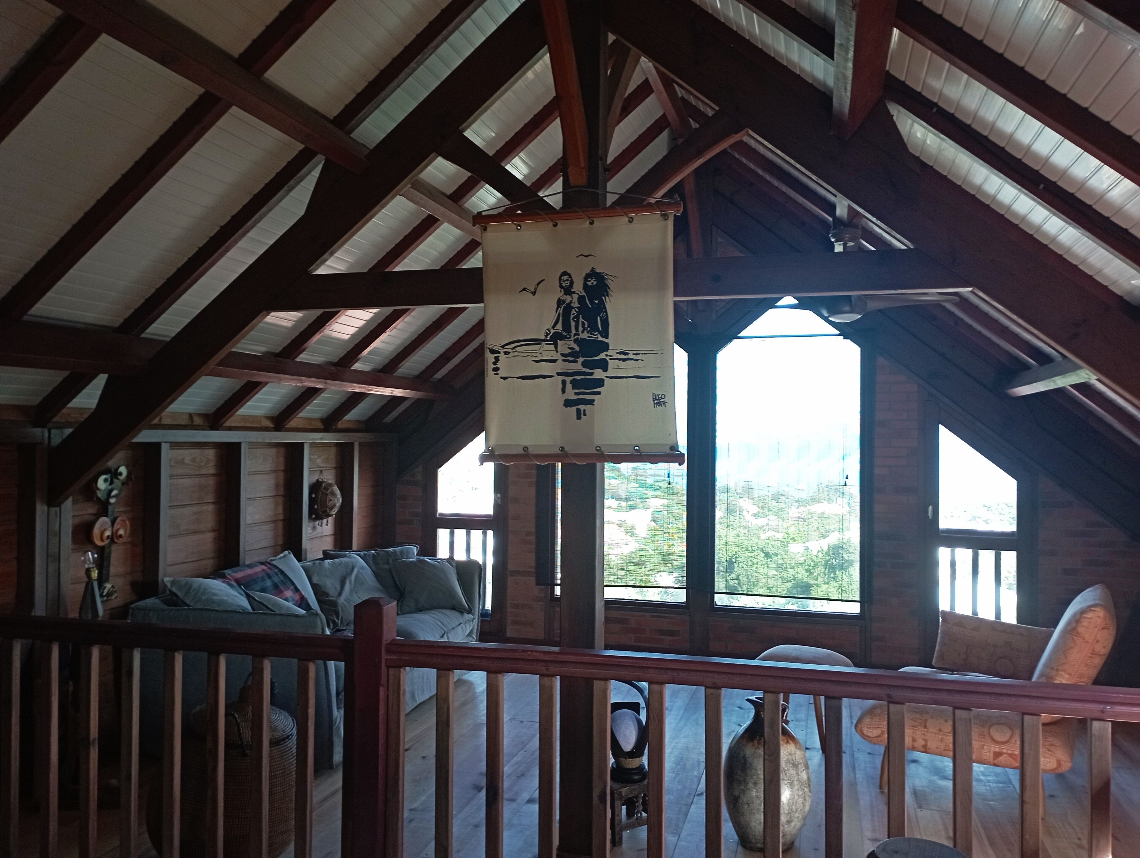 A rustic wooden interior with a high ceiling, a hanging sign, a sofa, and a view of greenery outside.