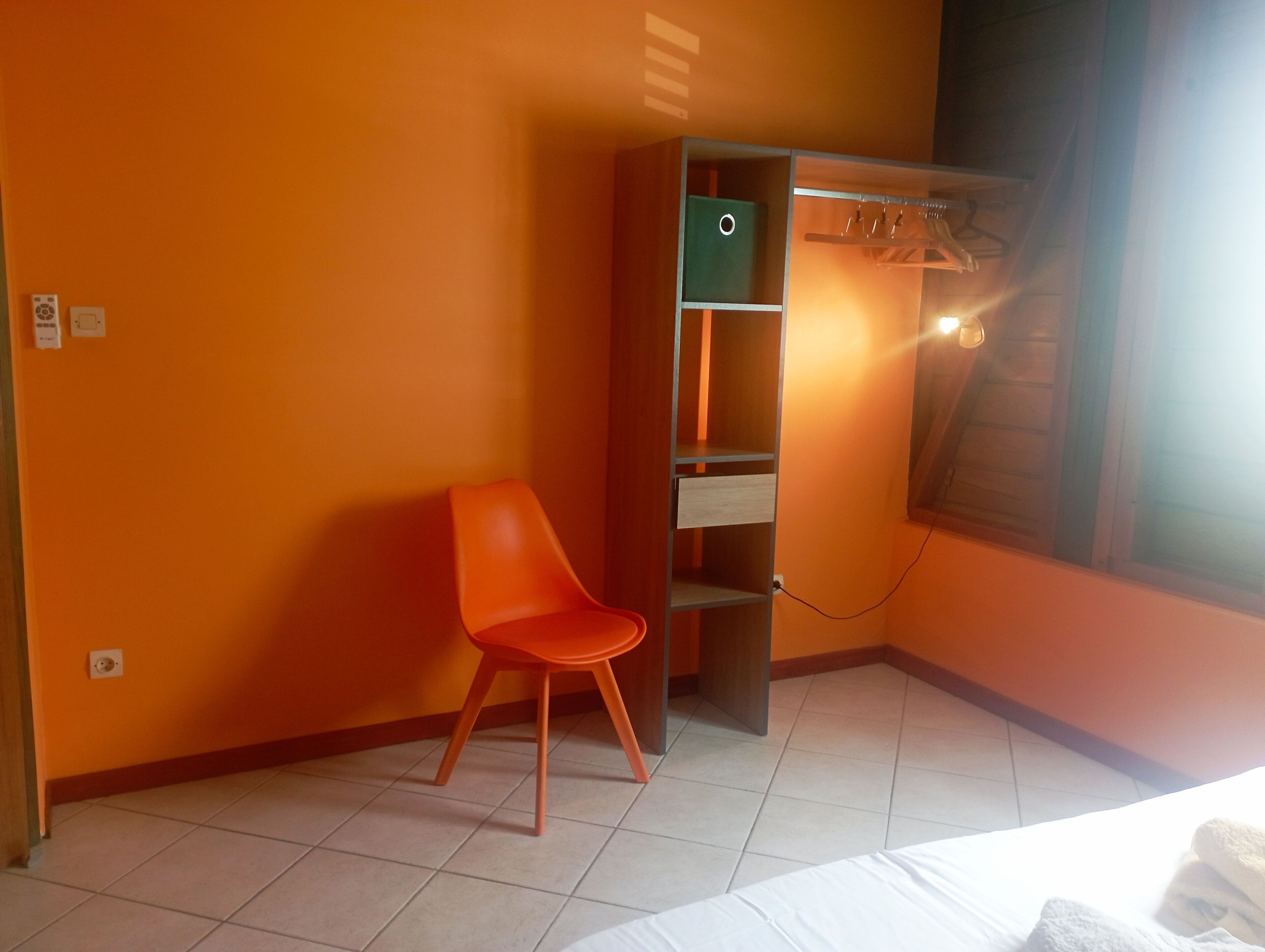 Ein Zimmer mit orangefarbenen Wänden, einem einzelnen orangefarbenen Sessel, einem Bett mit weißer Bettwäsche und einem Kleiderschrank mit Kleiderstange.