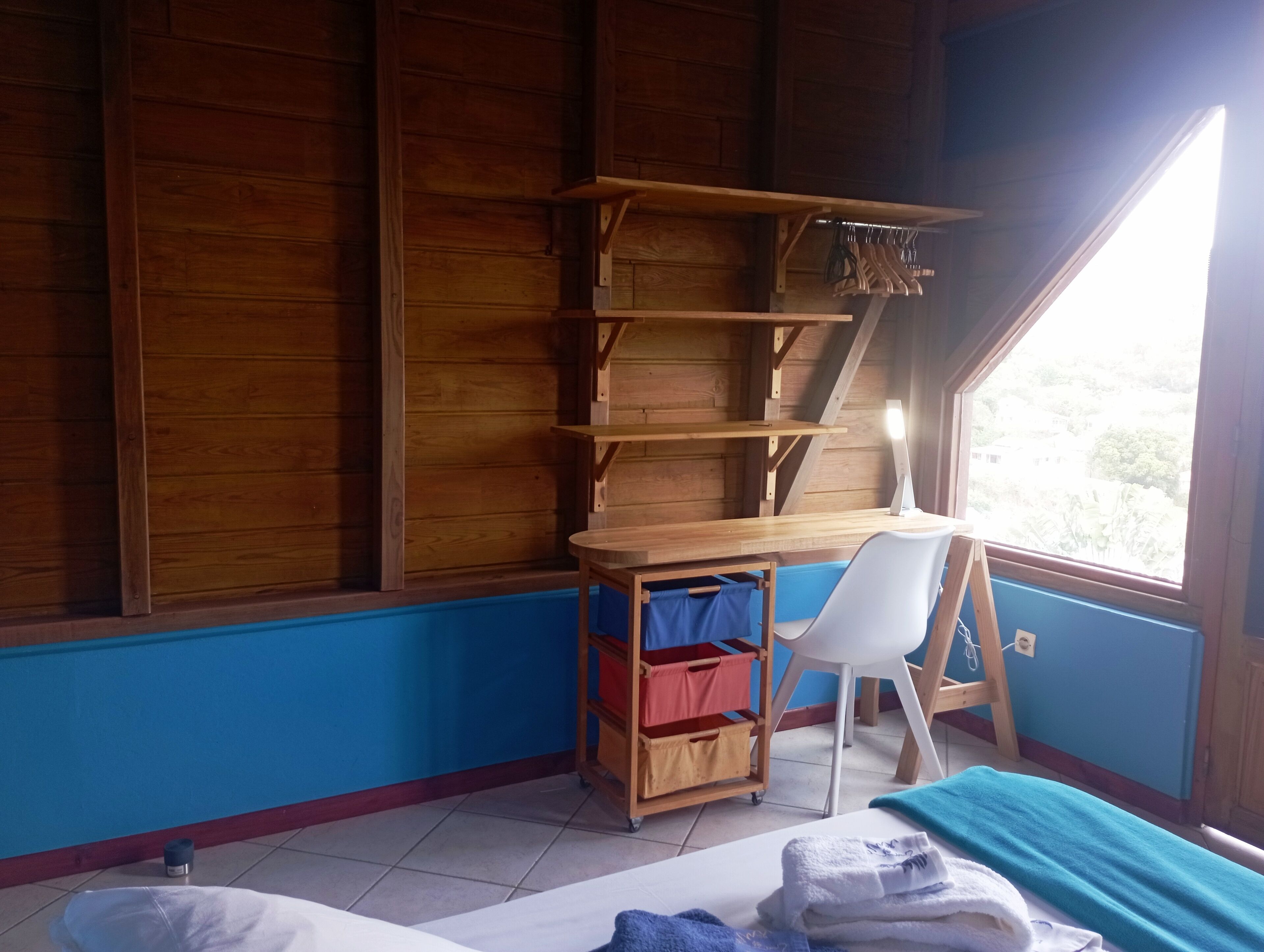 Ein Zimmer mit Holztisch, weißem Stuhl, blauer Wand und Fenster mit Blick auf Grünflächen.