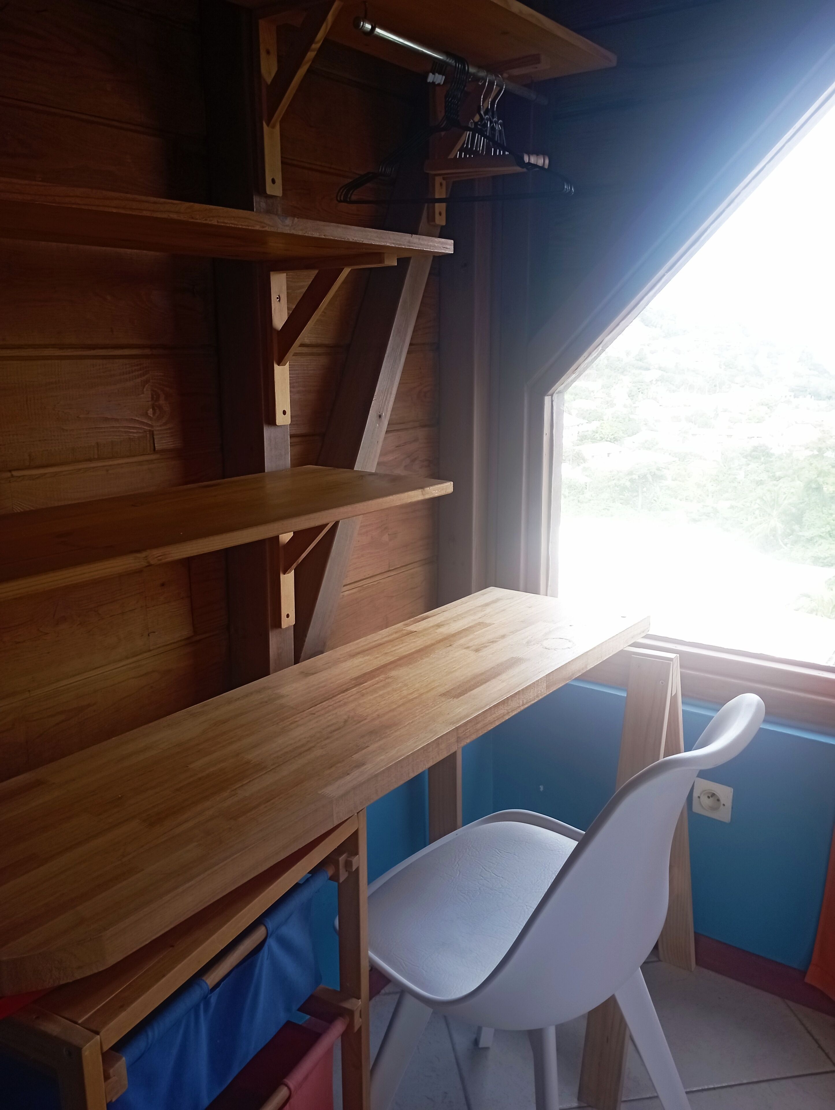Un escritorio de madera con una silla, una ventana con vistas a follaje verde, y una pared azul.