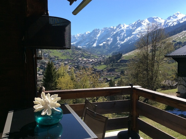 A balcony with a table and a vase of flowers, overlooking a mountainous landscape with snow-capped peaks and a valley below.
