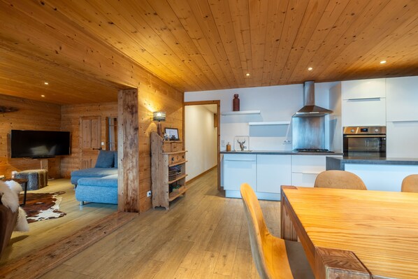 Un intérieur de chalet moderne avec un plafond en bois, un espace de vie comprenant un canapé et un téléviseur, une table à manger et une cuisine avec des armoires blanches et une cuisinière.