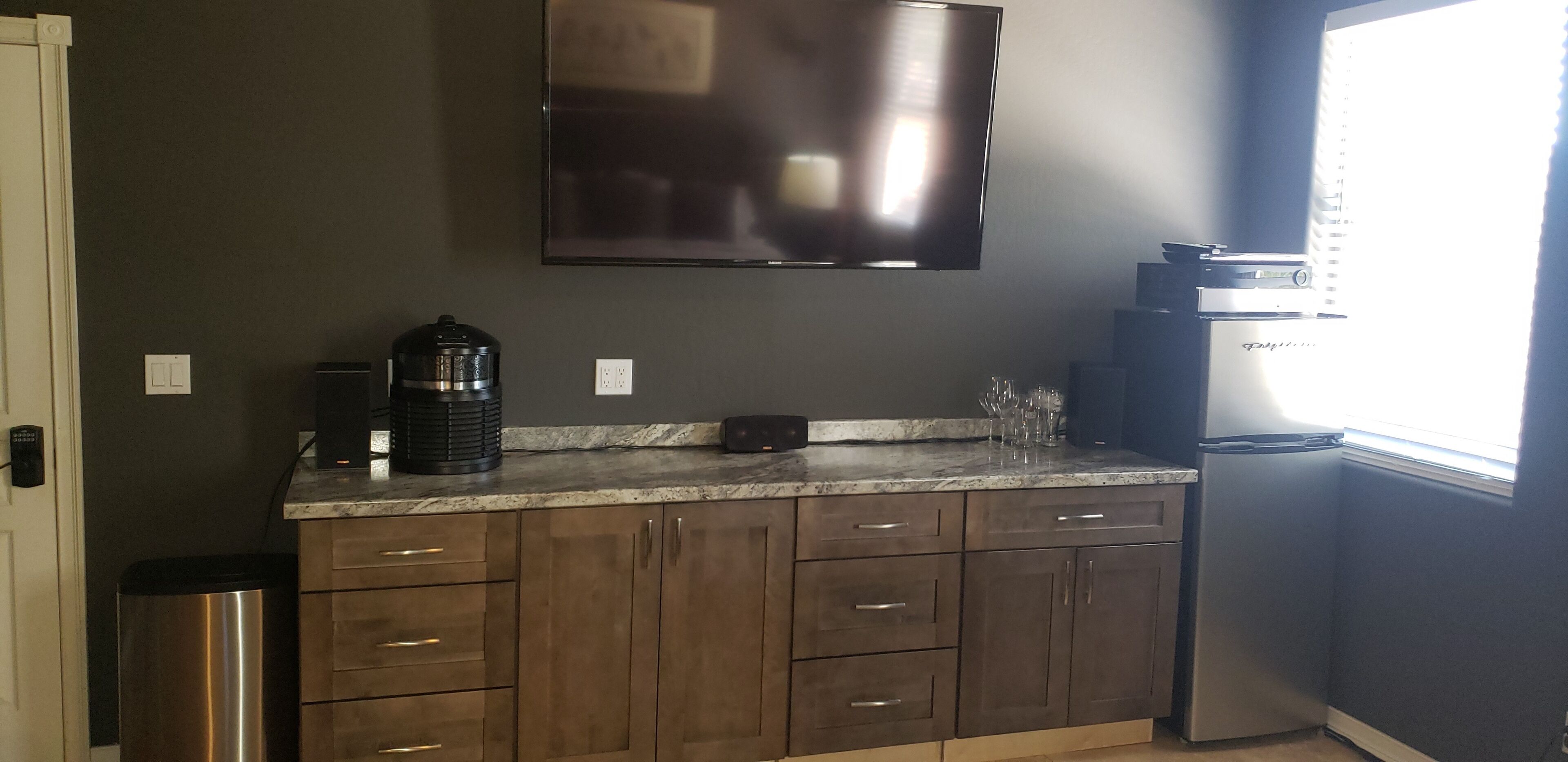 A kitchen with dark walls, a stainless steel refrigerator, a coffee maker, and a flat-screen TV mounted above the counter.