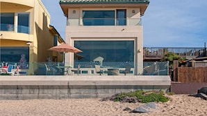 A beachfront property with a modern design, featuring a balcony with glass railings, a dining area with outdoor seating, and a view of the ocean.