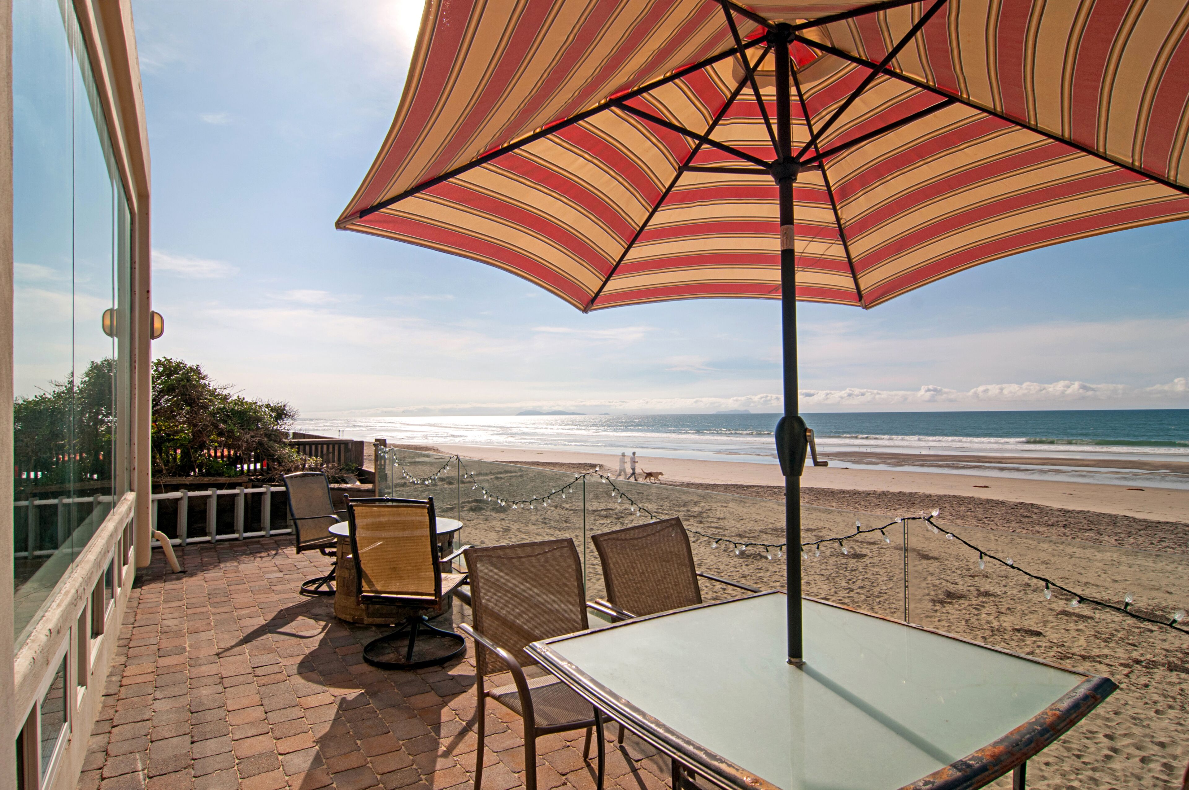 A patio with a table and chairs, overlooking a beach with a view of the ocean and distant mountains.