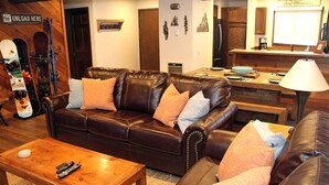 Un salon chaleureux avec un canapé en cuir, une table basse en bois et, au fond, un coin cuisine.