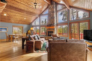 Amplia sala de estar con chimenea de piedra, techo de madera y ventanas grandes que dan a vistas de árboles.