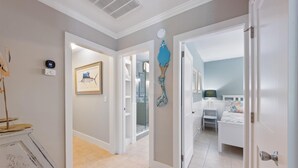 A hallway leading to a bedroom with a bed, a nightstand, and a dresser. There is a framed picture on the wall and a blue decorative item hanging on the door. - Willow Cottage: Steps to the beach, heated pool, perfect location! (Anna Maria)