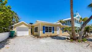 A two-story house with a garage, a covered porch, and a small garden area with palm trees. - Willow Cottage: Steps to the beach, heated pool, perfect location! (Anna Maria)