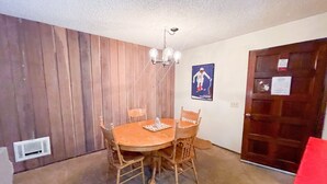 Een eetkamer met een houten tafel en stoelen, een kroonluchter, een poster aan de muur en een deur met een bericht.