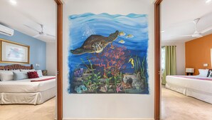 Habitación de hotel con un mural en la pared que muestra una tortuga marina y peces, una cama, un sofá y un ventilador de techo.