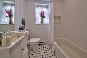 A bathroom with a toilet, sink, and bathtub. There are two windows with curtains, and a small shelf above the toilet.