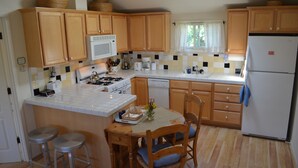 A compact kitchen with wooden cabinets, a white refrigerator, a microwave, and a gas stove. There are two stools and a table with a vase of flowers. - Sweet 1 BDRM Cottage, FAST WIFI, near ocean, W&D, Full kitchen and bath, BBQ! (Santa Cruz)
