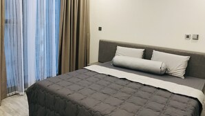 Una habitación de hotel moderna con una cama grande, un cabecero gris y una ventana con cortinas translúcidas.