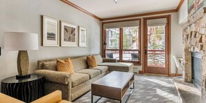 A living room with a sofa, coffee table, and a fireplace. There are framed pictures on the wall and a window with a view of trees.