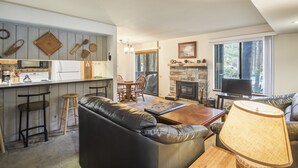 A modern kitchen with bar stools, a dining area with a wooden table and chairs, a fireplace, and a living room with a leather sofa and a lamp. - Sunshine Village 167 (Mammoth Lakes)