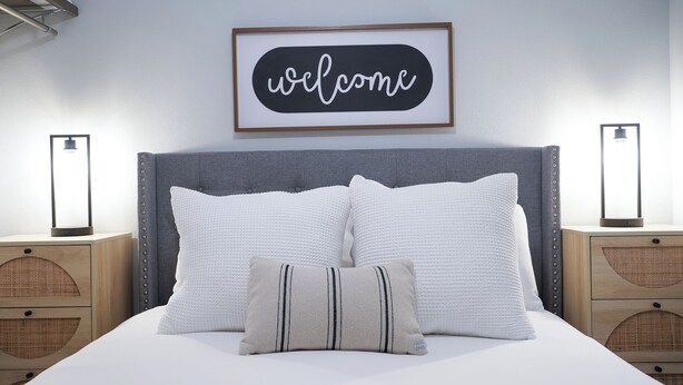 Un lit bien fait, avec une literie blanche, une tête de lit rembourrée de couleur grise et un panneau encadré avec l’inscription « Bienvenue » placé au-dessus.