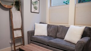 Un canapé gris avec des coussins blancs, une échelle en bois, une plante en pot et une fenêtre avec des stores.