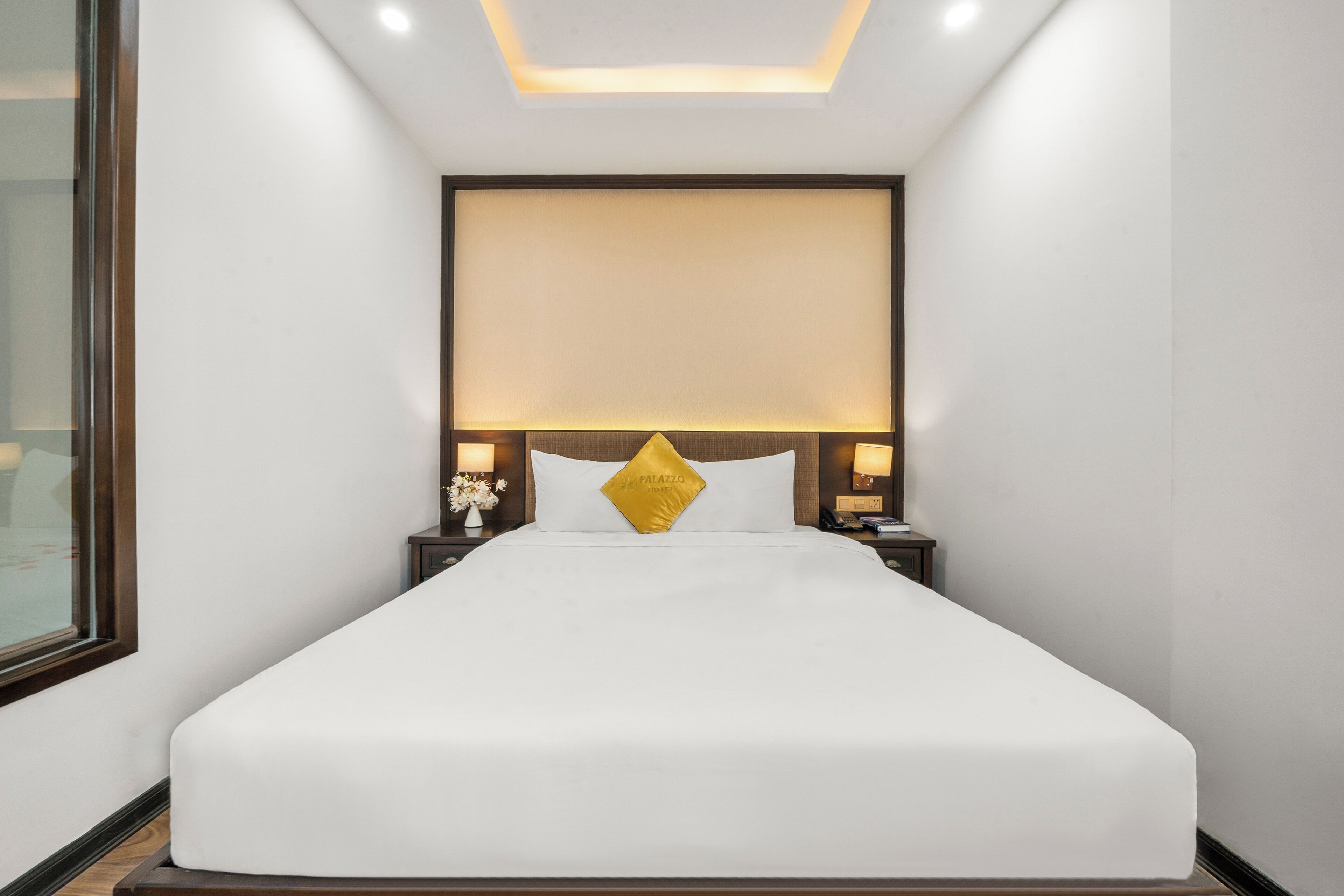 A neatly made bed with a white bedspread and a gold accent pillow, flanked by two bedside tables with lamps, in a modern hotel room with a recessed ceiling light.