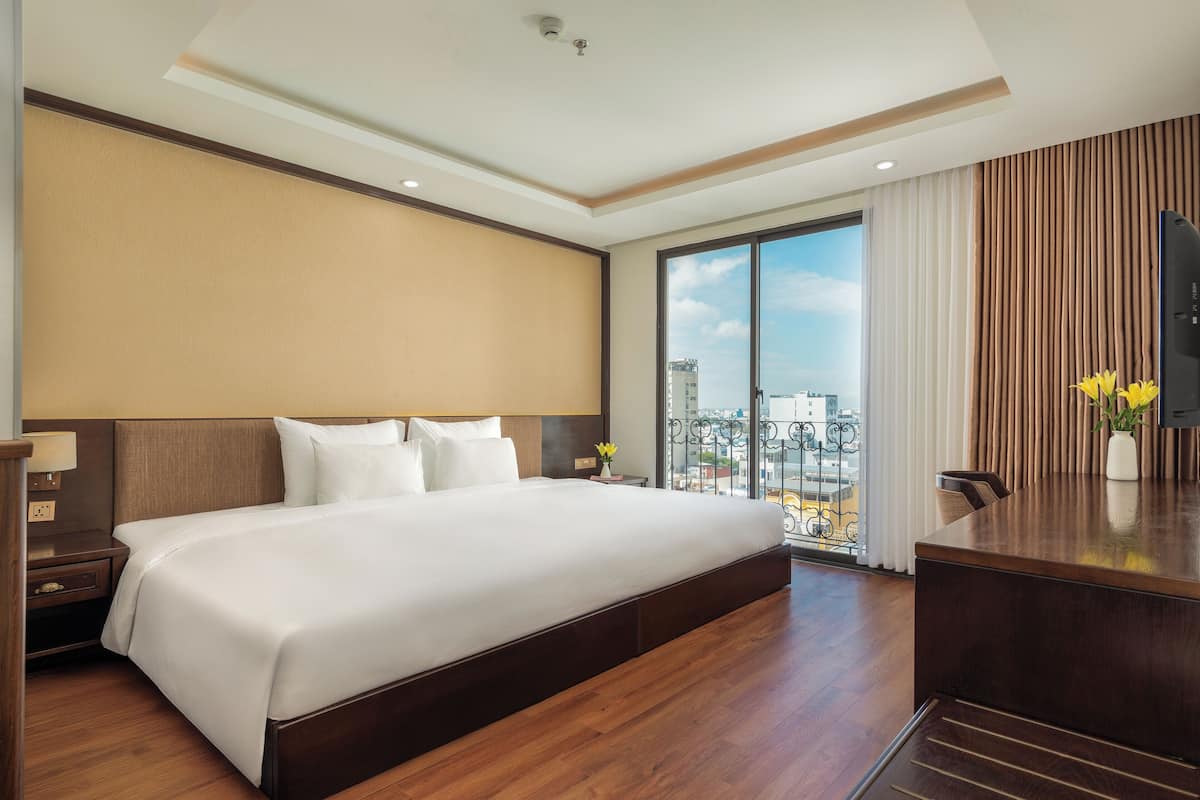 A hotel room with a large bed, a desk, and a view of the city through large windows.