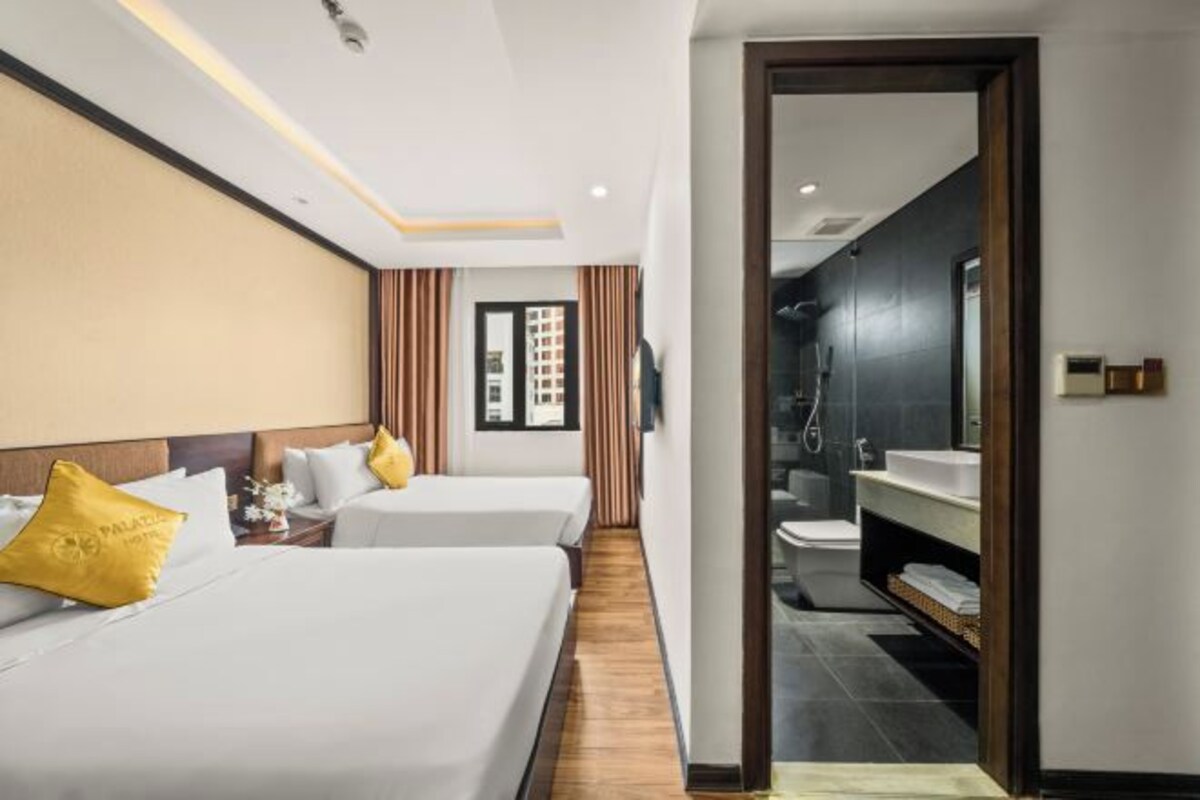 A hotel room with two beds, a wooden floor, a bathroom with a sink and mirror, and a view of the city.