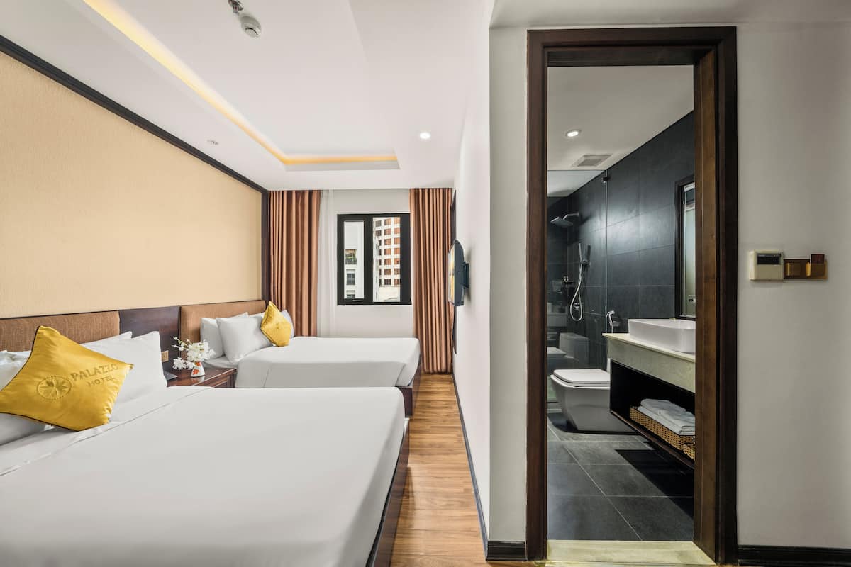 A hotel room with two beds, a wooden floor, a bathroom with a sink and mirror, and a view of a building outside.