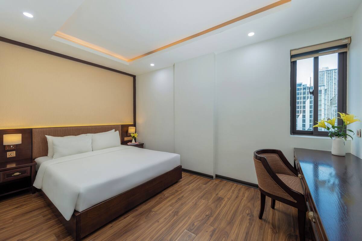 A hotel room with a large bed, wooden flooring, a desk with a chair, a window with a view of buildings, and a nightstand with a lamp.