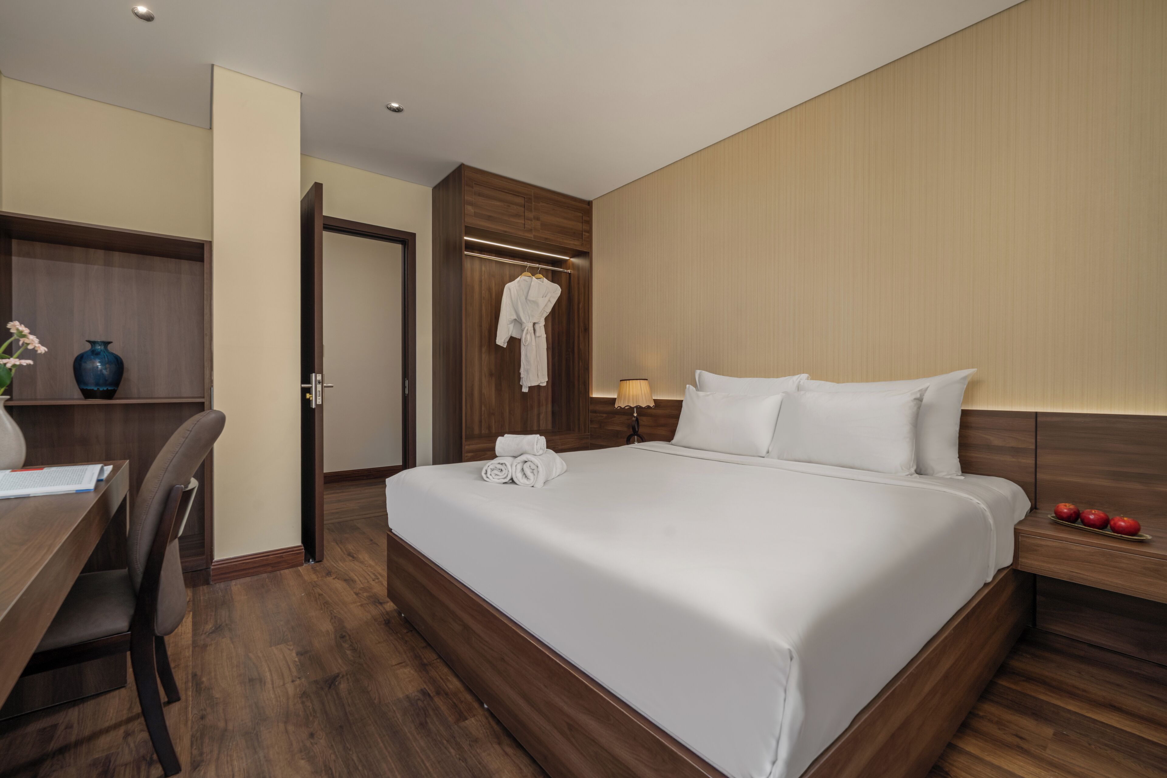 A hotel room with a large bed, a desk, a chair, a wardrobe, and a bedside table with a lamp and apples.