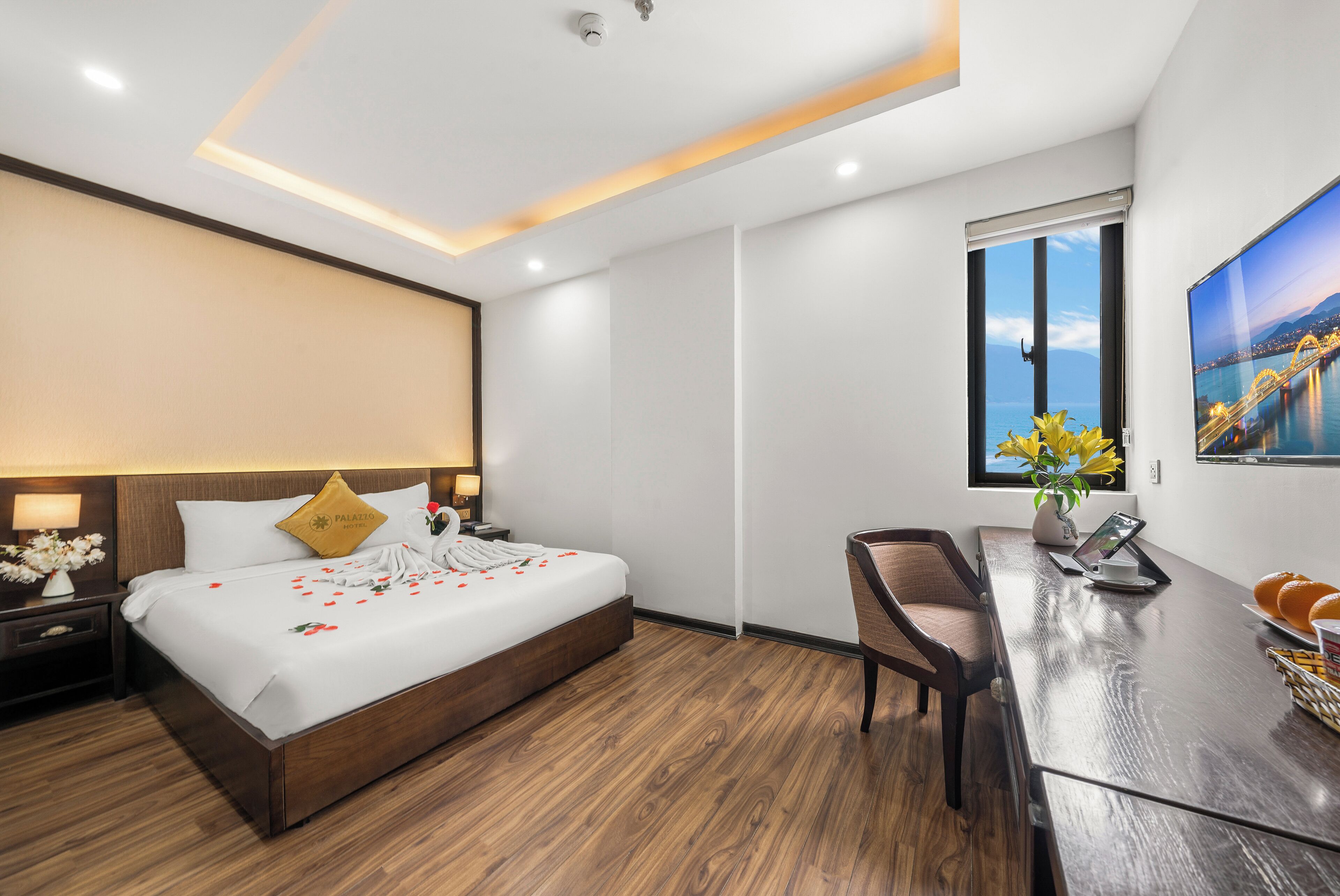 Una habitación de hotel moderna con suelo de madera, una cama grande con ropa blanca, un escritorio con silla, una ventana con vistas a la ciudad y un cuadro en la pared.