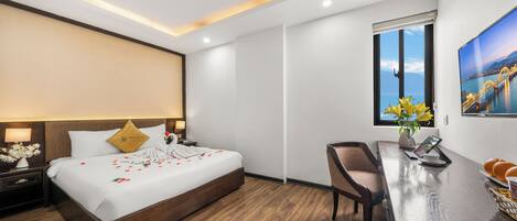Una habitación de hotel moderna con suelo de madera, una cama grande con ropa blanca, un escritorio con silla, una ventana con vistas a la ciudad y un cuadro en la pared.