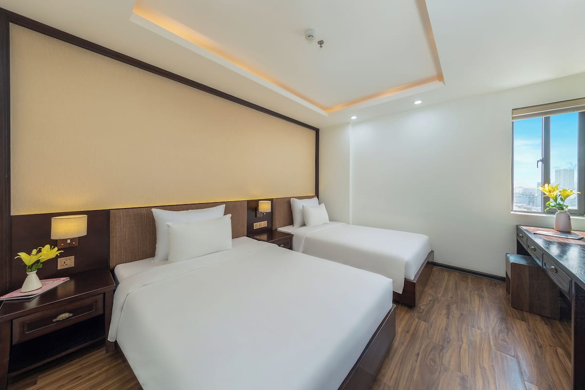 A hotel room with two beds, a desk, a nightstand, a lamp, and a window with a city view.