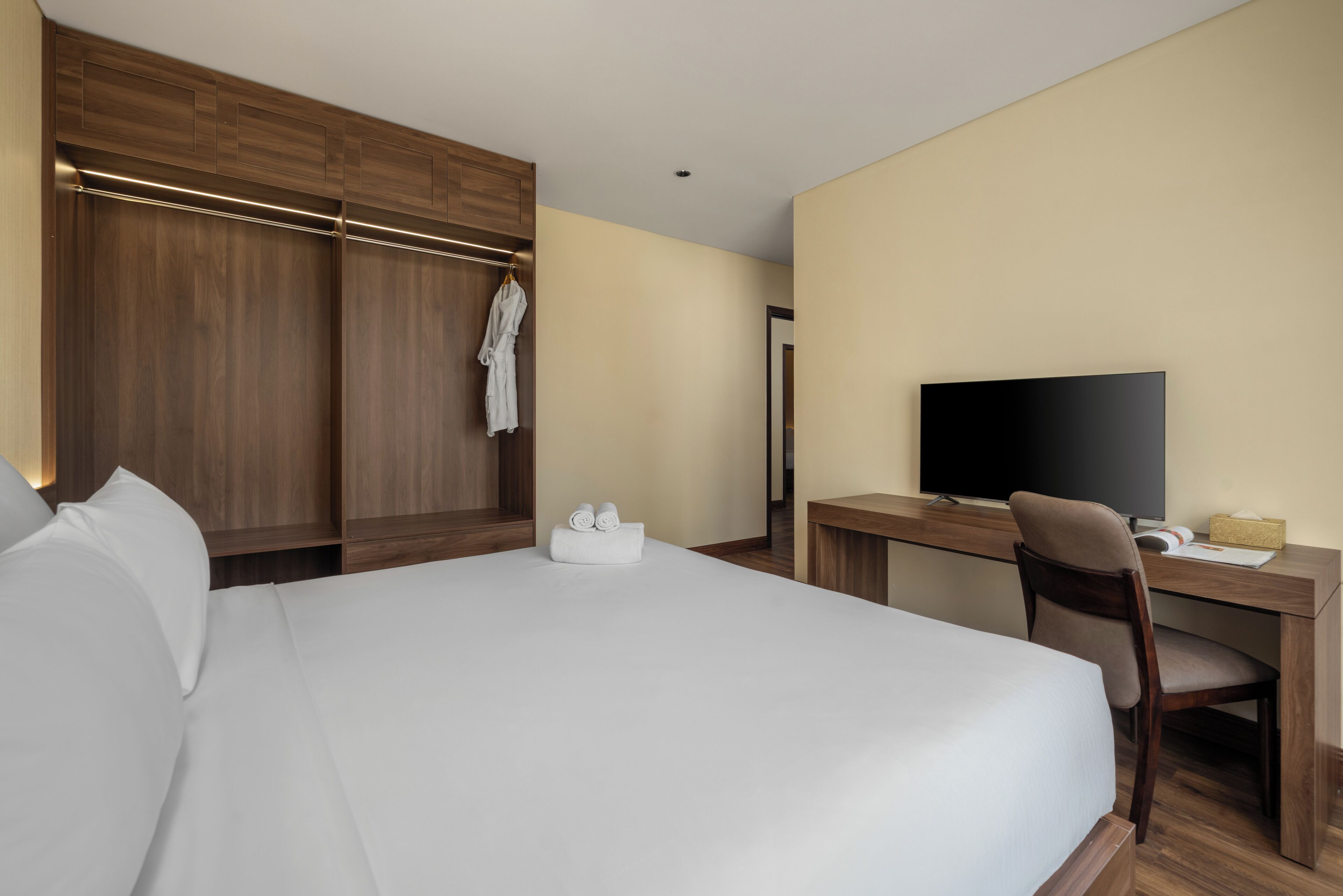 A hotel room with a large bed, wooden wardrobe, a desk with a chair, a television, and a bathroom visible through an open door.