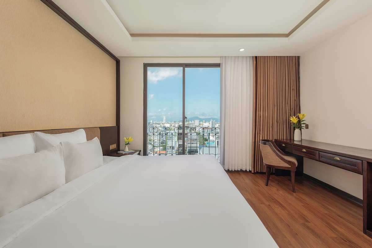 A hotel room with a large bed, a desk, and a view of a cityscape through a large window.