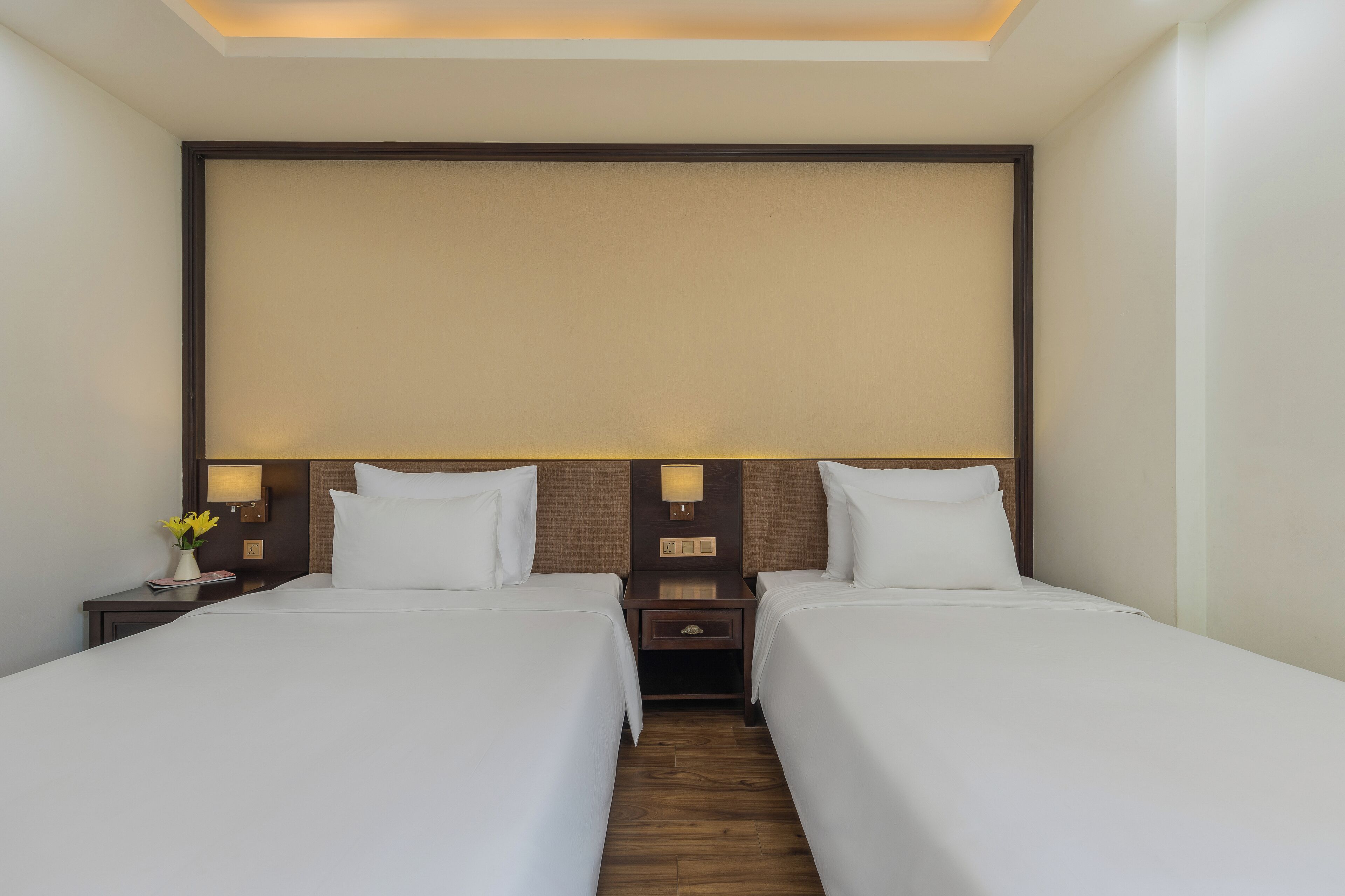 A hotel room with two single beds, a nightstand with a lamp, and a wall-mounted panel.