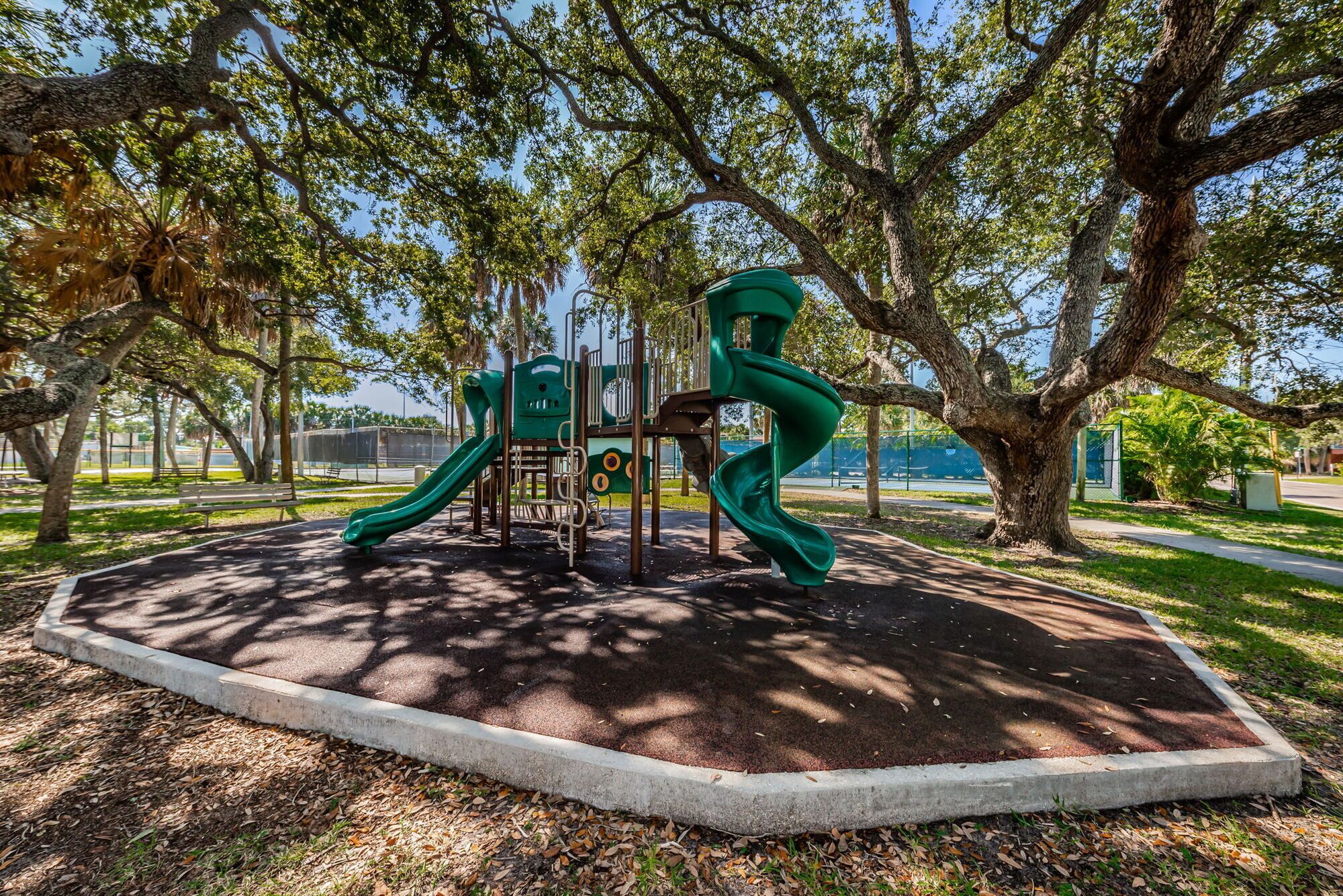 A playground with a slide and climbing structures, surrounded by trees and a paved path.