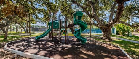 A playground with a slide and climbing structures, surrounded by trees and a paved path.