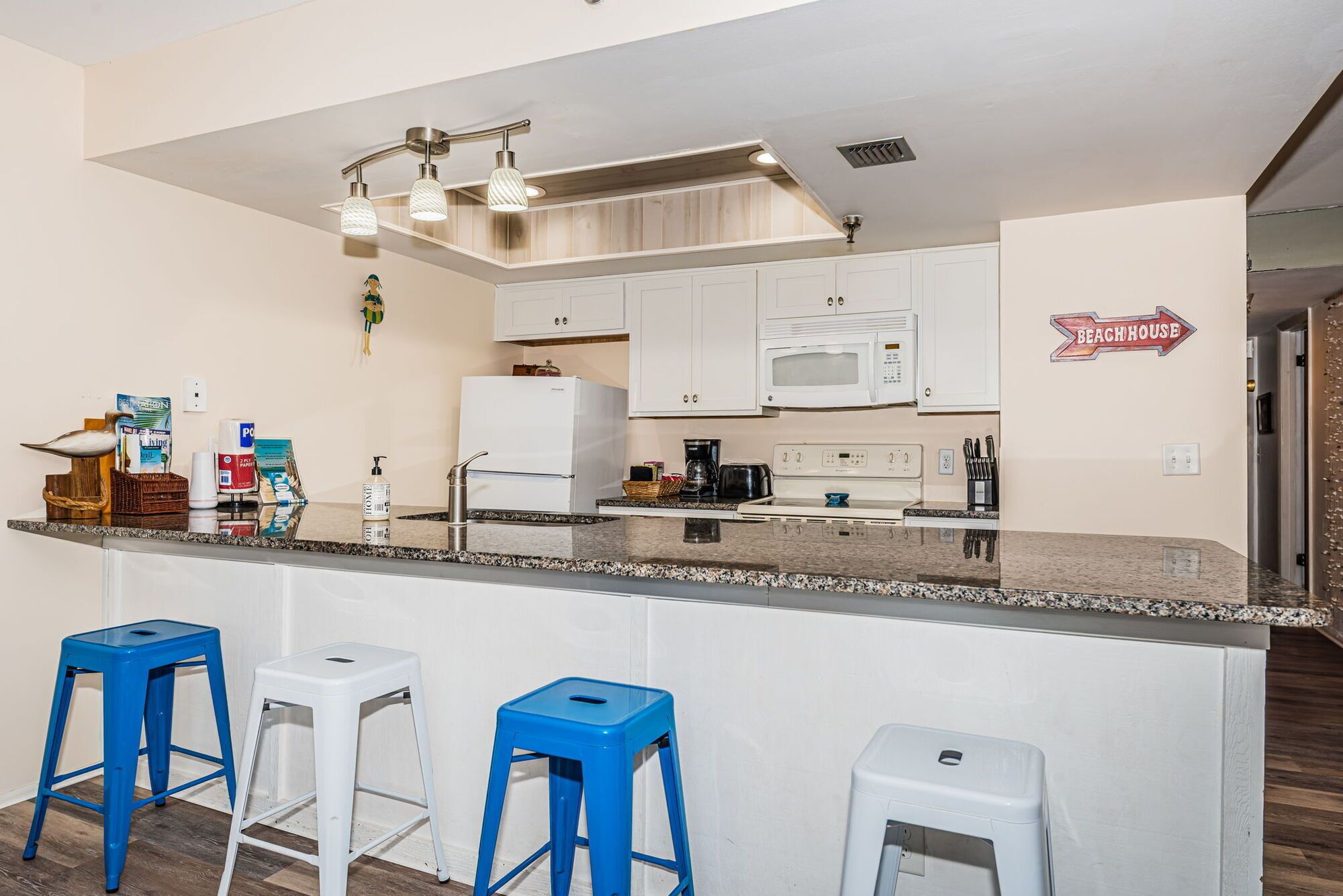 Una cocina con armarios blancos, un microondas, una cafetera y un letrero que dice 'Beachhouse'. Hay tres taburetes azules y un refrigerador blanco.